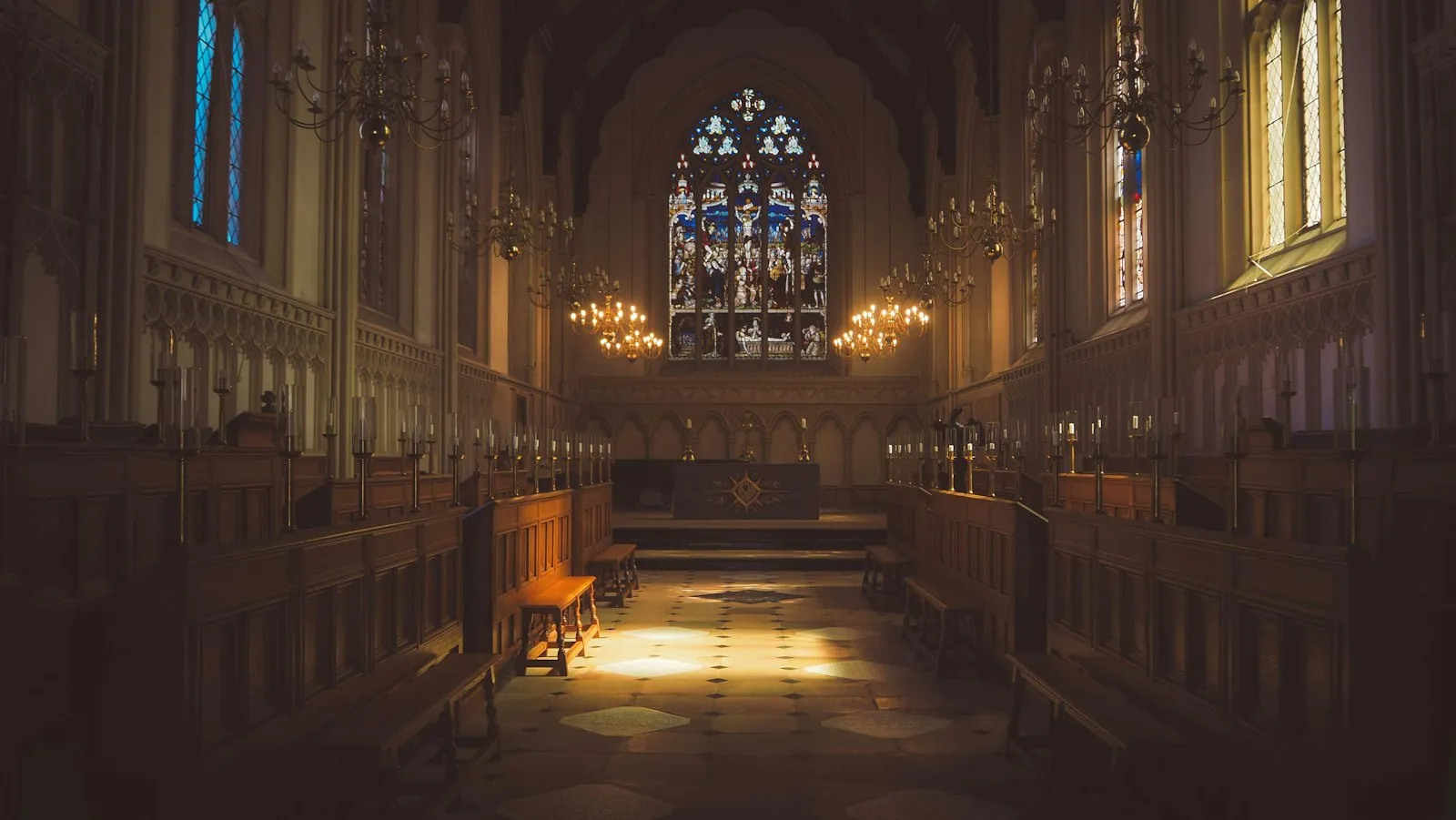 The interior of a Gothic chapel with carved wooden pews, chandeliers and a stained glass window
