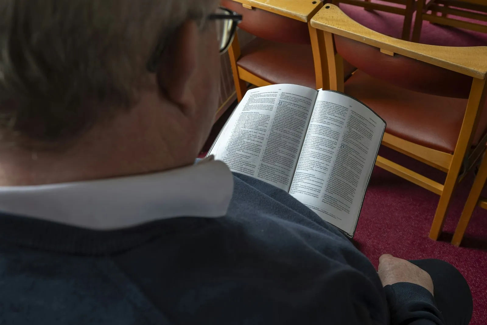 An older man in a sweater reading an open Bible, seen over his shoulder
