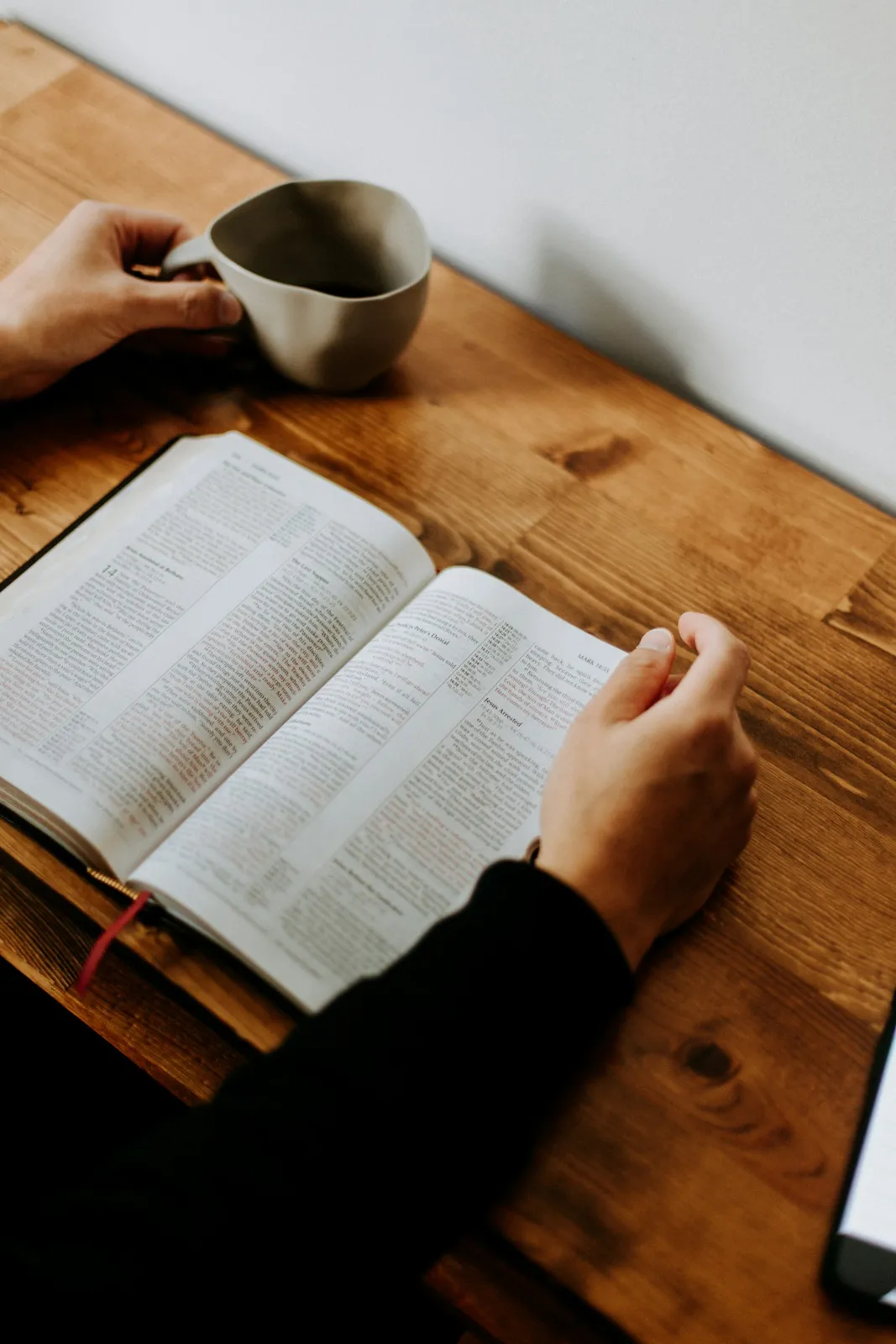 An open Bible on a wooden table beside a hand holding a coffee mug